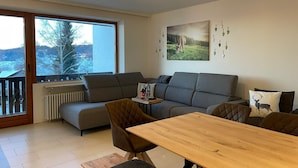 Un salon moderne avec un canapé, une table à manger en bois et une vue sur un paysage enneigé, visible à travers de grandes fenêtres.