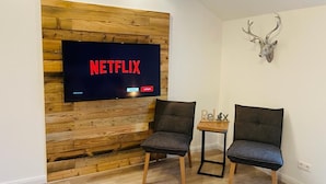 Un salon moderne avec un mur recouvert de panneaux en bois, un téléviseur fixé au mur affichant le logo « NETFLIX », deux fauteuils sombres, une petite table sur laquelle est posée une pancarte « Relax » et une décoration représentant une tête de cerf.