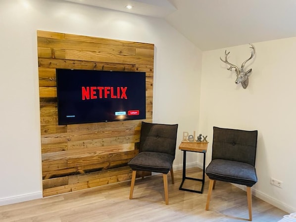 Ett modernt vardagsrum med en träpanel på väggen, en väggmonterad tv som visar "NETFLIX", två mörka fåtöljer, ett litet bord med en skylt som säger "Avkoppling" och en dekorativ hjorthuvudtrofé.