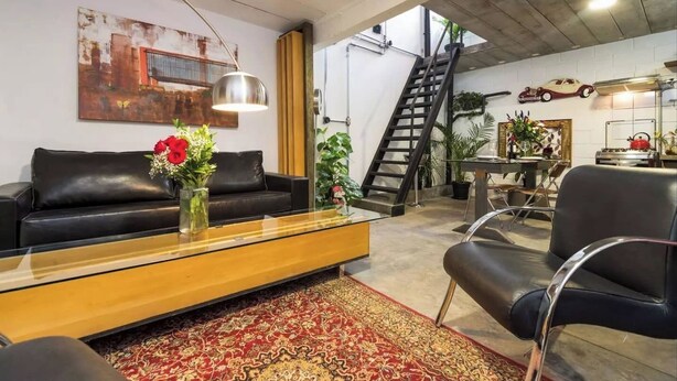 Ein modernes Wohnzimmer mit einem schwarzen Ledersofa, einem Holztisch und einem roten Teppich mit Muster. Eine Treppe führt zu einer oberen Ebene, und es gibt einen Essbereich mit Tisch und Stühlen.