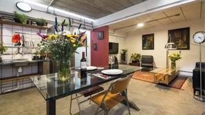 Eine moderne Küche mit einem Glastisch, Stühlen und einem Blumenstrauß. Ein Fernseher ist an der Wand montiert und eine Uhr hängt dort.