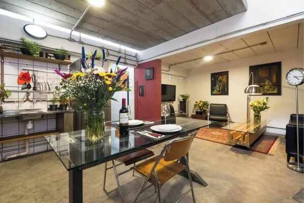 A modern kitchen with a glass dining table, chairs, and a vase of flowers. There is a TV mounted on the wall and a clock.