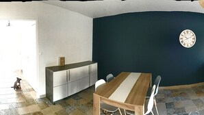 A room with a dark blue accent wall, a wooden dining table with chairs, a grey sofa, a TV, and a small dog in the doorway. - 77m2 T4 apartment (La Seyne-sur-Mer)