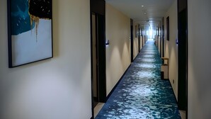 Un long couloir étroit, recouvert d’un tapis à motifs bleus et blancs, avec des œuvres d’art encadrées accrochées au mur gauche, et des portes de part et d’autre.