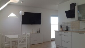 Une cuisine moderne avec des meubles blancs, une table à manger avec des chaises, un téléviseur fixé au mur et une fenêtre laissant entrer la lumière naturelle.