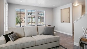 Un salon avec un canapé beige, un tapis à motifs et une vue sur un immeuble par de grandes fenêtres.