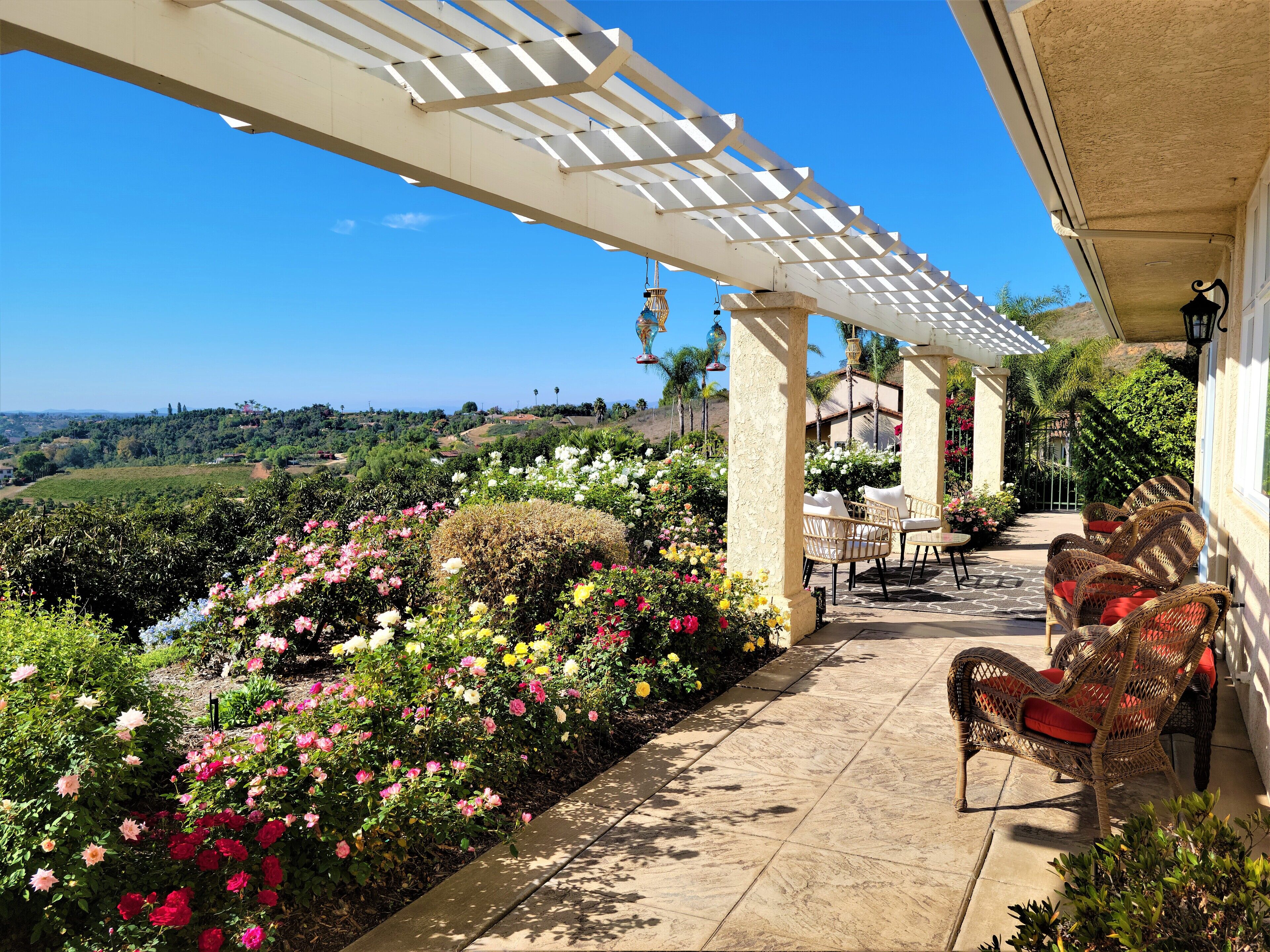 Un patio cubierto con mobiliario exterior, una pérgola y vistas a un jardín ajardinado y colinas al lejano.