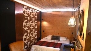 Ein gemütliches Zimmer mit einem Bett, einem Sessel, einer Holzwand mit Kuhfellmuster, einer Pendelleuchte und einem kleinen Waschbecken mit Pflegeprodukten.