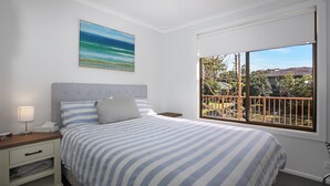 A bedroom with a bed, a nightstand, a bedside lamp, a window with a view of the outside, and a painting on the wall. - Barralong Beach House (Hawks Nest)