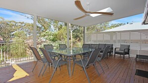 An outdoor patio with a glass table and chairs, a ceiling fan, and a view of trees and houses. - Barralong Beach House (Hawks Nest)