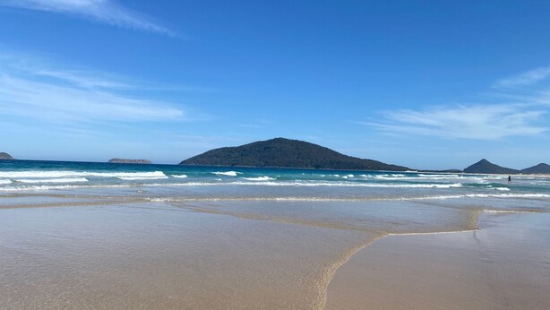 A beach with clear blue water, sandy shore, and distant hills. - Barralong Beach House (Hawks Nest)