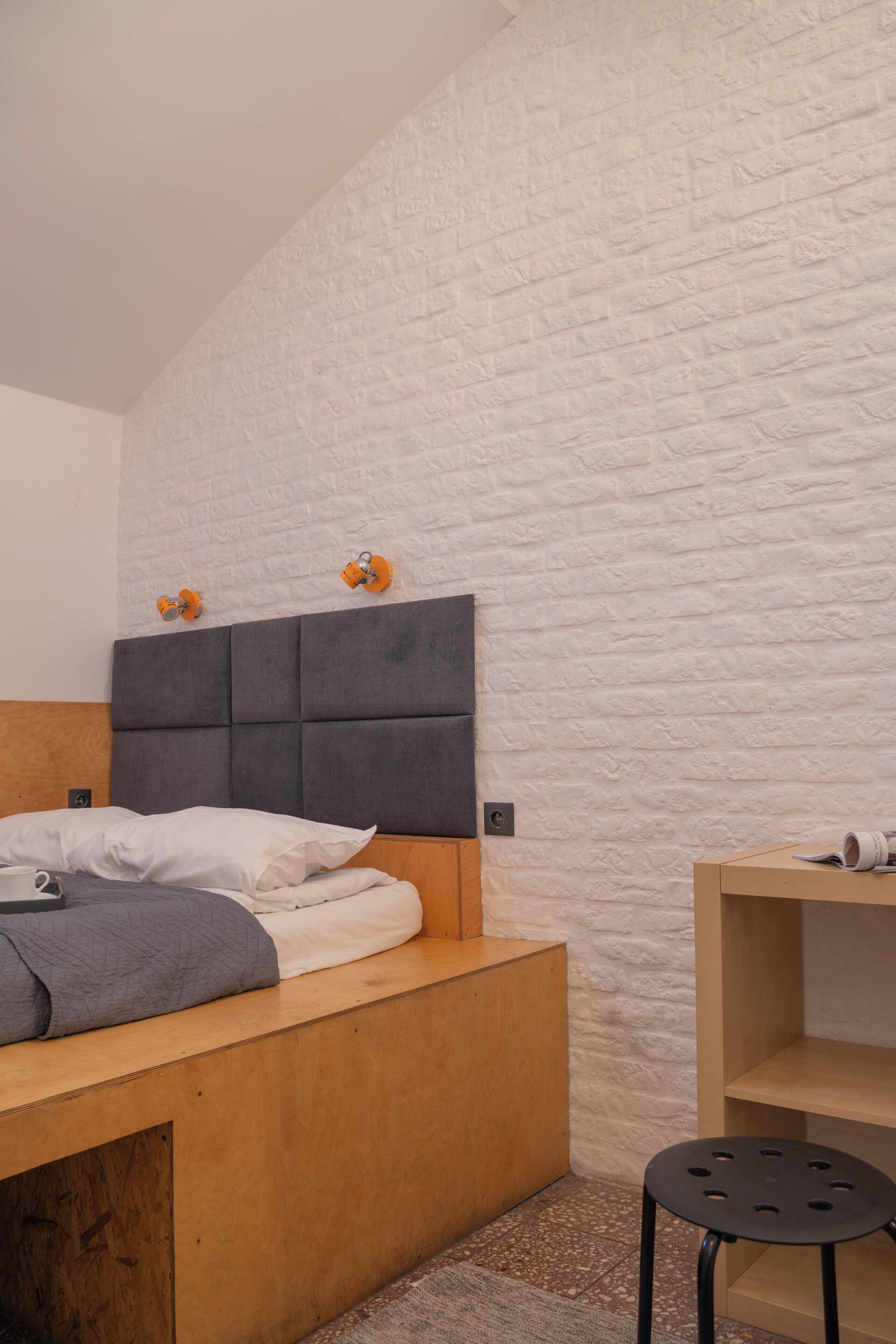 A bedroom with a wooden bed, a grey headboard, a white brick wall, and a small wooden nightstand.