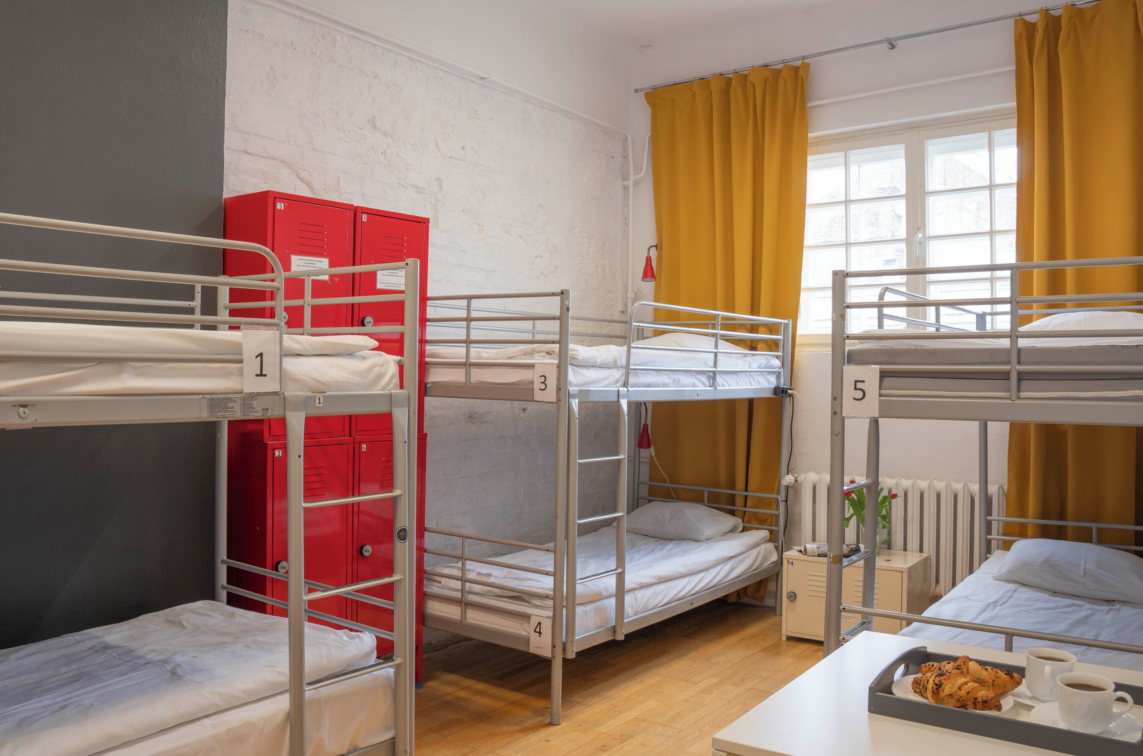 Ein Schlafsaalzimmer mit Stockbetten, nummerierten Schließfächern, einem Heizkörper und einer Platte mit Essen und Getränken.