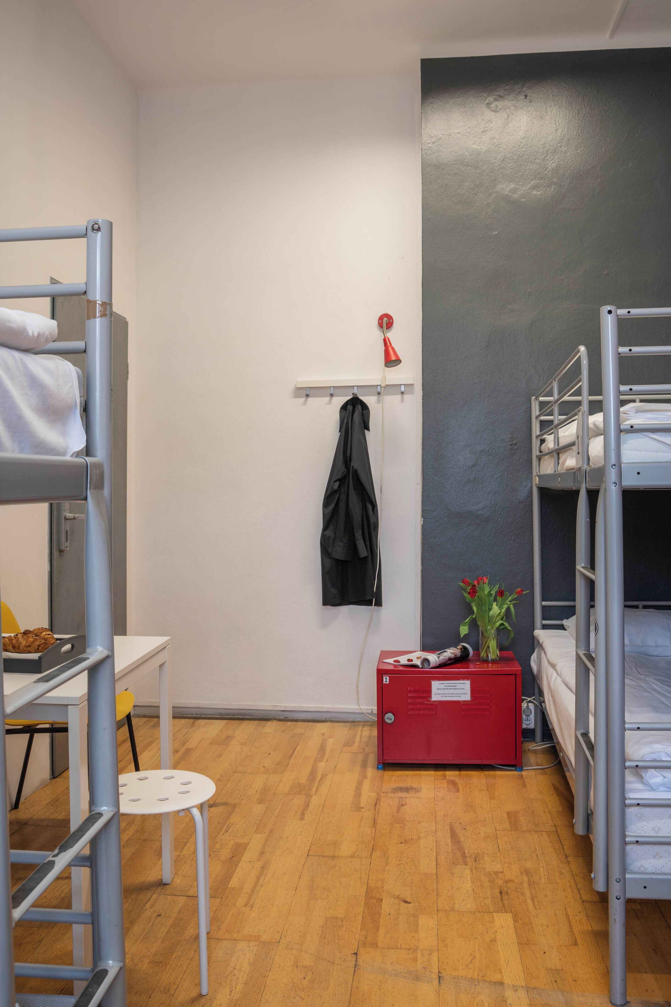 A dormitory room with bunk beds, a small table, a chair, a shelf with a jacket, and a red storage unit with flowers.
