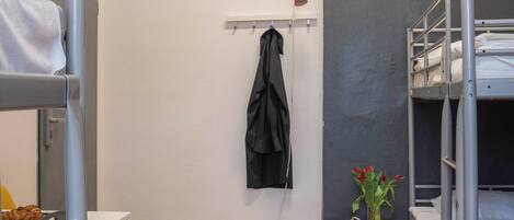 A dormitory room with bunk beds, a small table, a chair, a shelf with a jacket, and a red storage unit with flowers.
