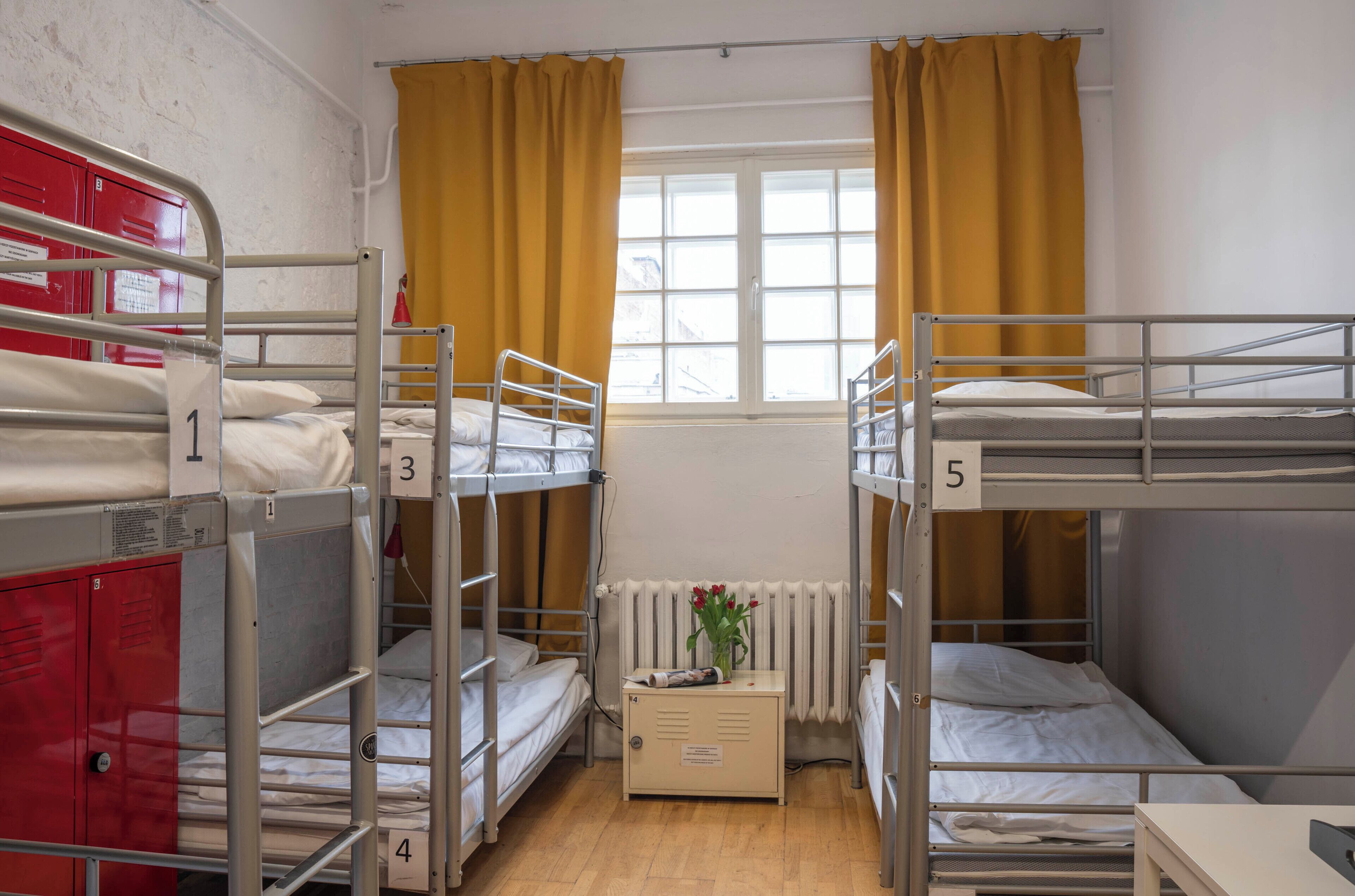 A dormitory room with bunk beds, numbered lockers, a radiator, and a window with curtains.