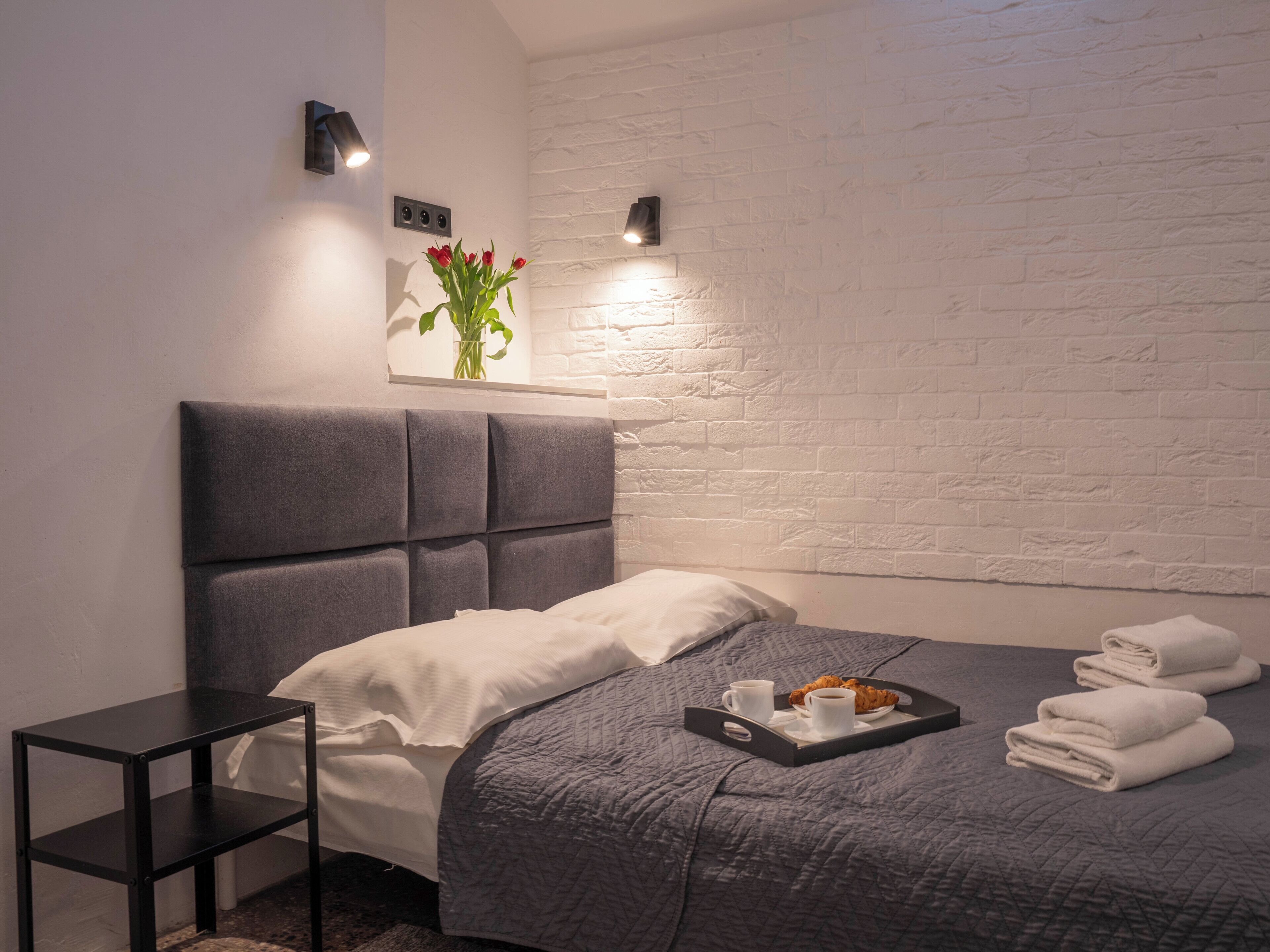 Ein modernes Schlafzimmer mit grauem Kopfteil, einem Nachttisch, einer Ablage mit Handtüchern und einer Speiseplatte, sowie einer Wandleuchte.