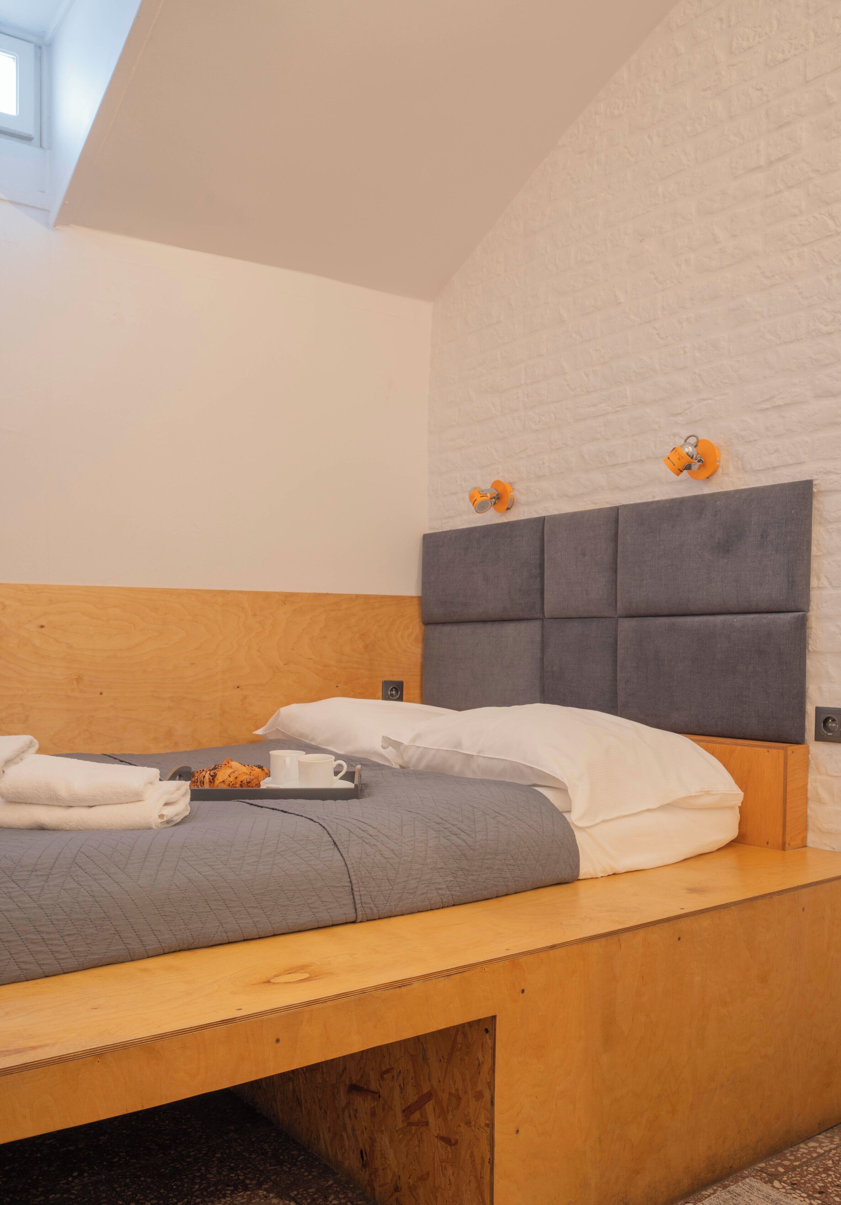 A modern bedroom with a wooden headboard, a grey quilted bedspread, and a tray with towels and a cup.