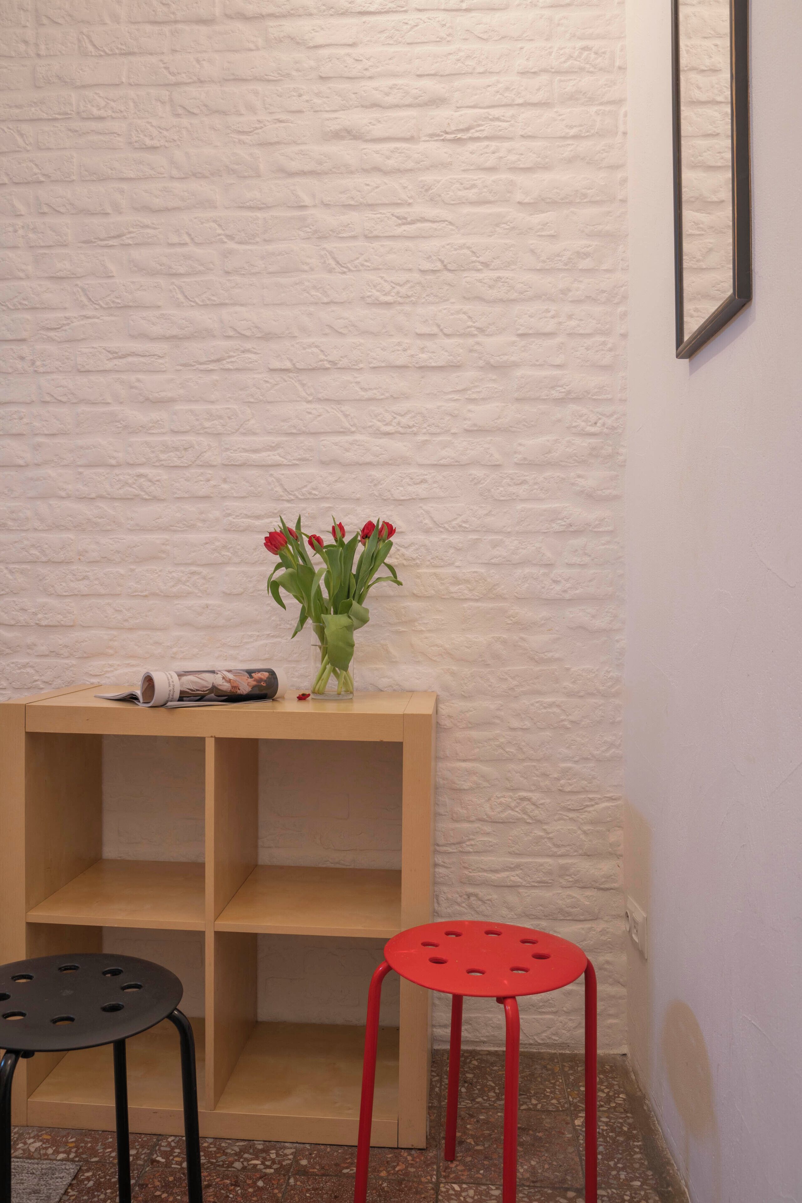 A corner with a white brick wall, a wooden shelf with a magazine, a vase with red flowers, and two stools.