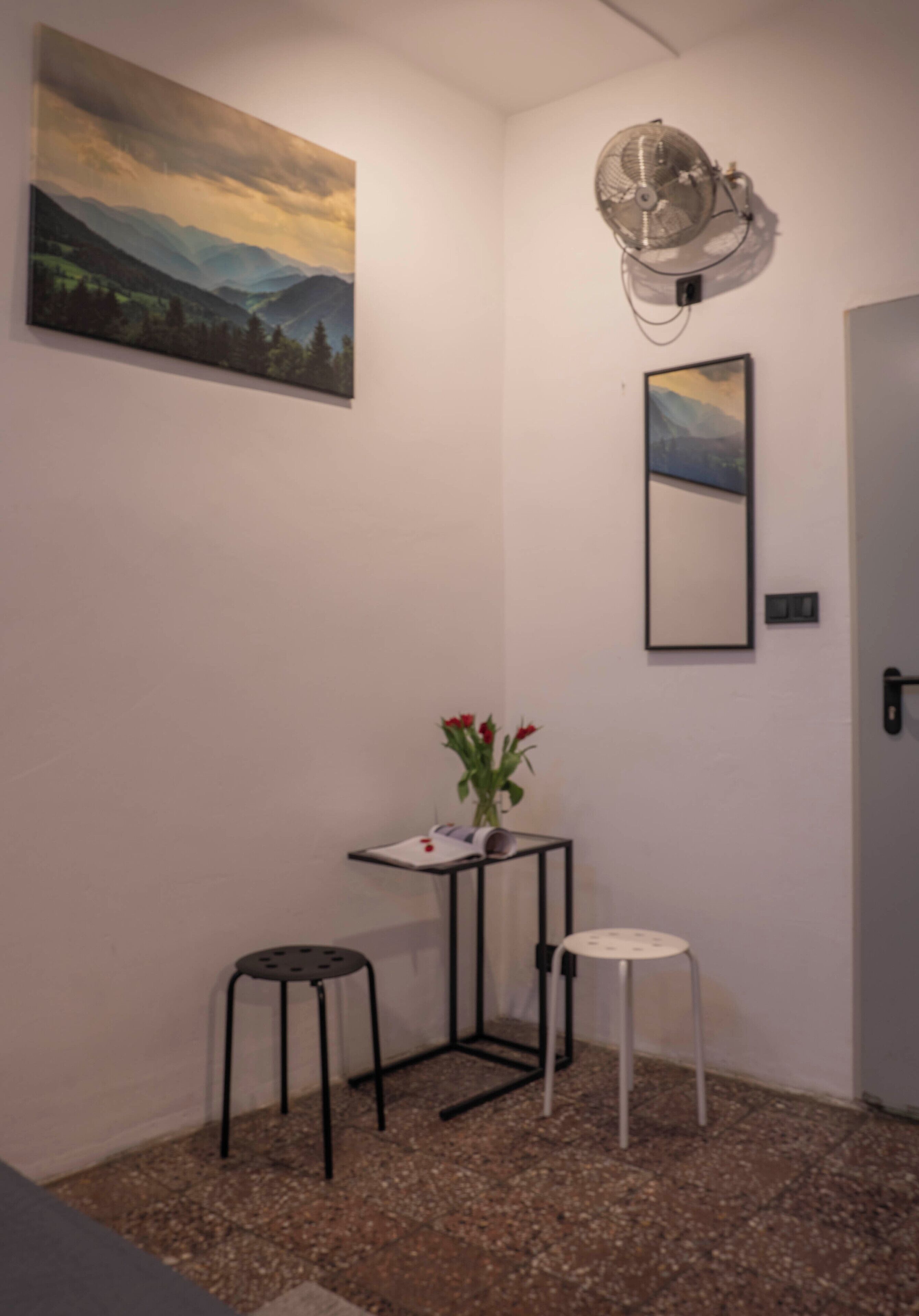 Una habitación pequeña con un cuadro de un paisaje montañoso, un ventilador, un espejo y una mesita con un florero.