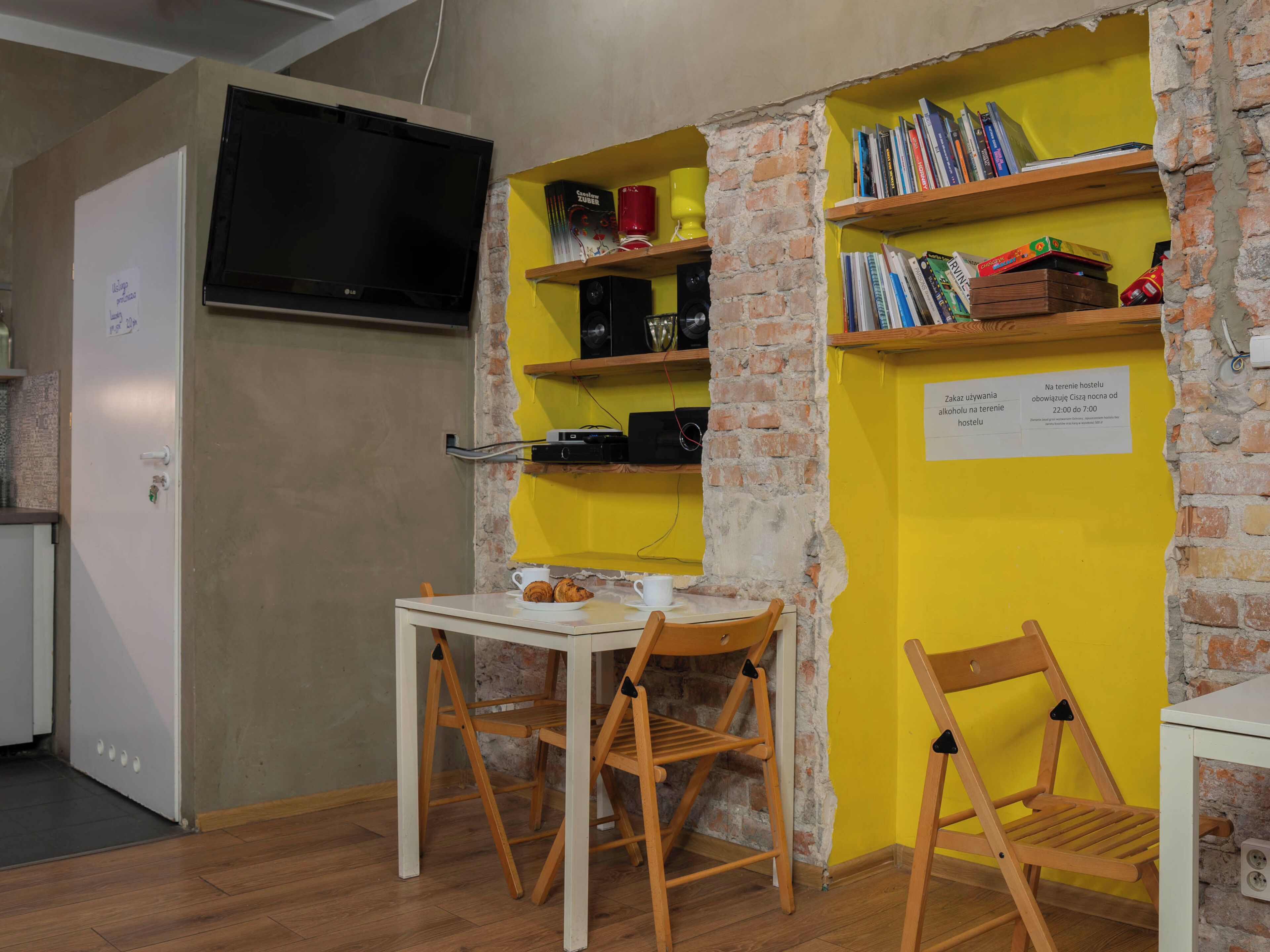 A room with a brick wall, a television mounted above a white cabinet, a table with chairs, and shelves with books and decorative items.