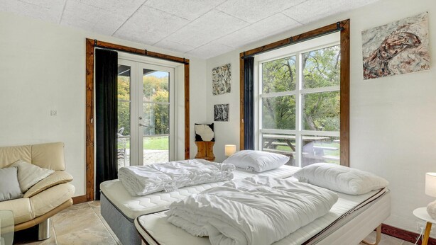 A bedroom with a bed, a chair, and a view of the outdoors through large windows.