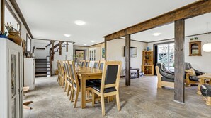 A spacious dining area with a long wooden table and chairs, a staircase, and a living area with a sofa and a bookshelf.