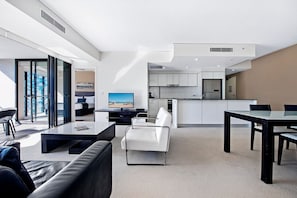 A modern living space with a sofa, a dining table, a television, and a kitchen area. - Stunning 2 Bed Apartment in Sierra Grand (Broadbeach)