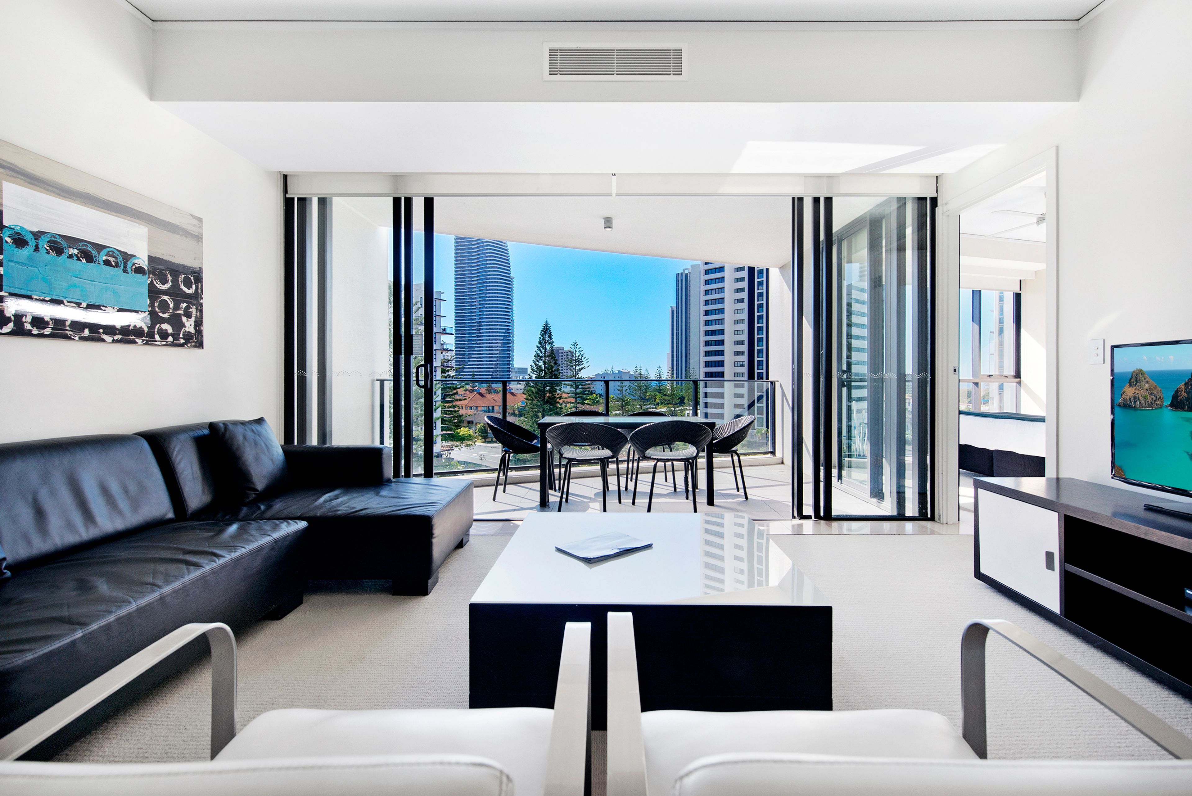 A modern living room with a sofa, a dining table, and chairs, a flat-screen TV, and a balcony with a city view.