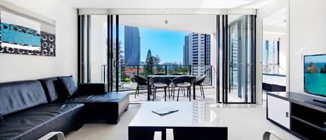 Una sala de estar moderna con sofá, mesa de comedor, sillas, televisión de pantalla plana y balcón con vistas a la ciudad.