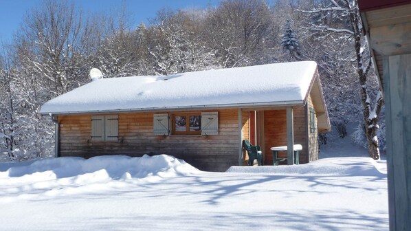 A wooden cabin with a snow-covered roof, a porch with two chairs, and a small table.