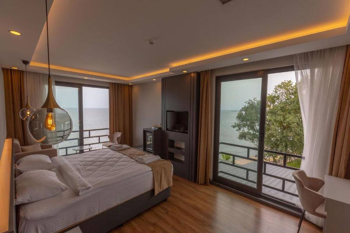 A modern bedroom with a large bed, a TV, and a balcony with a view of the sea.