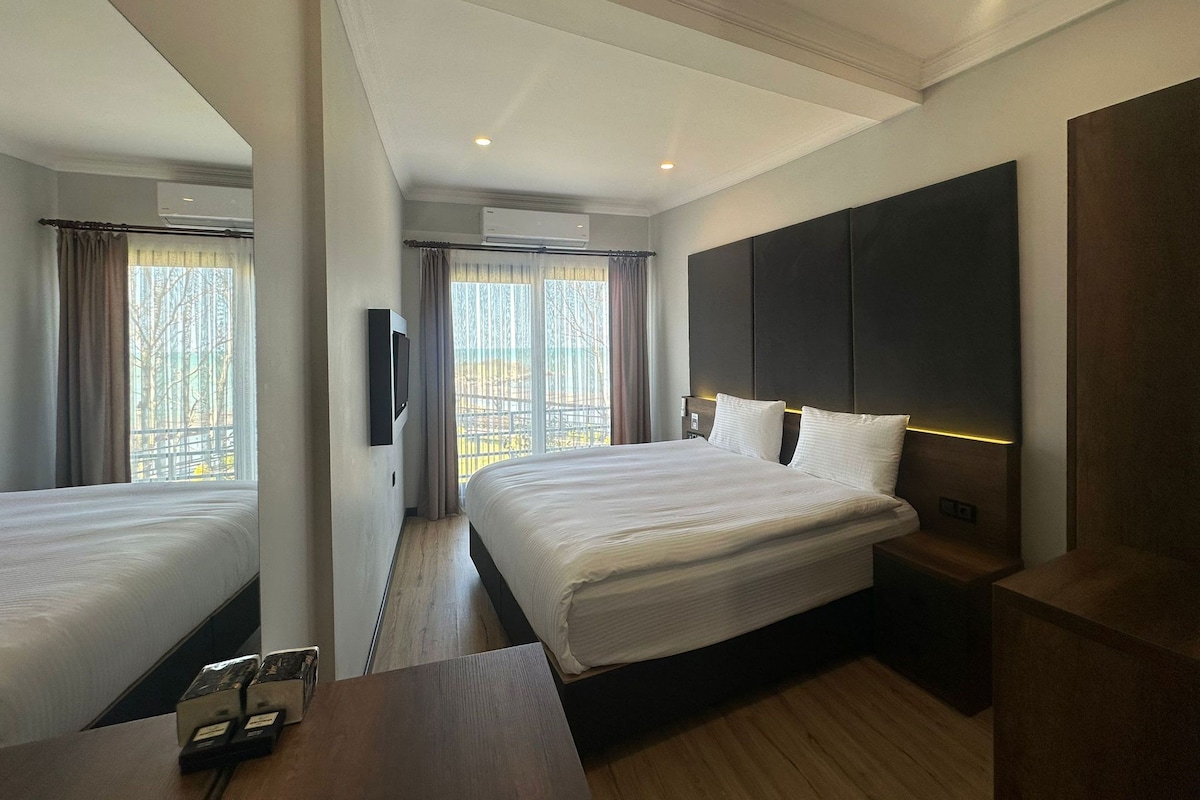 A hotel room with a large bed, a bedside table, a TV mounted on the wall, and a window with curtains.