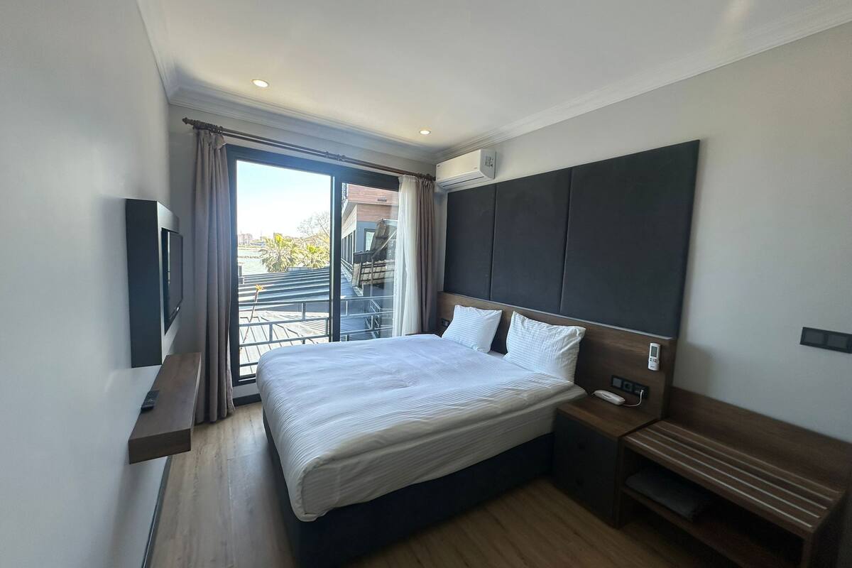 A modern hotel room with a large bed, a TV, a bench, and a view of the outdoors.