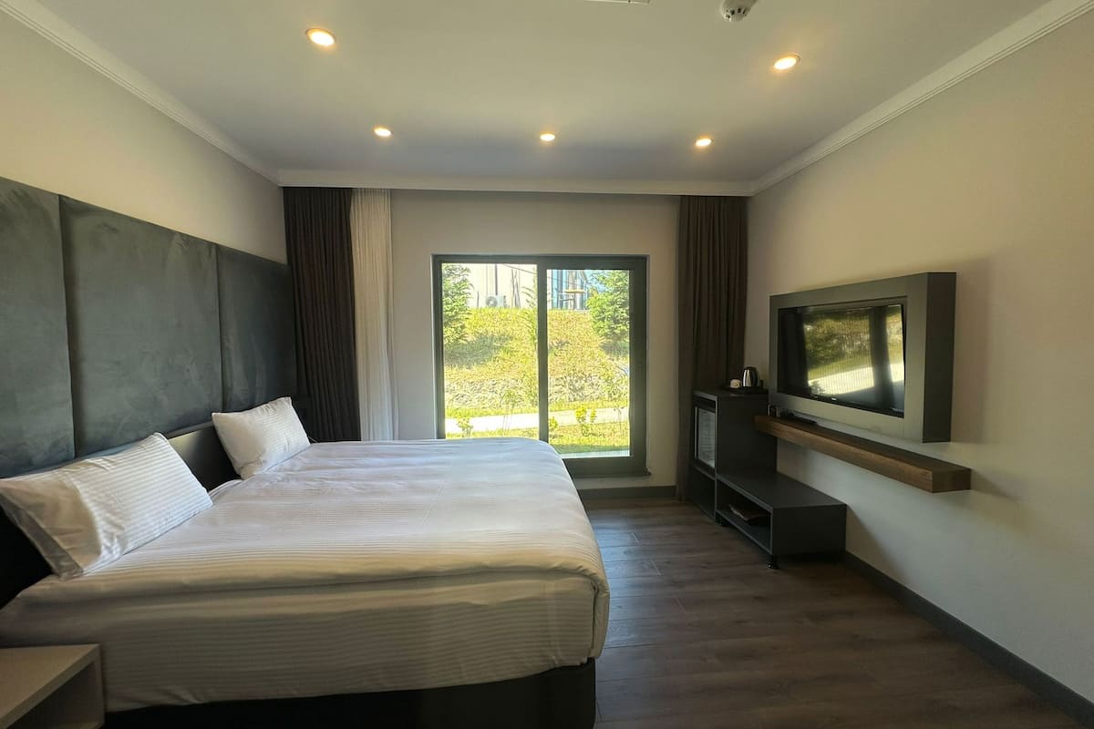 A modern hotel room with a large bed, a flat-screen TV, and a view of greenery outside.