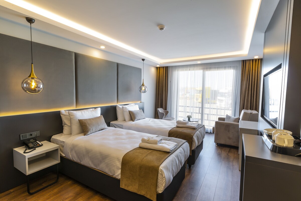Habitación de hotel con dos camas, un escritorio, una silla y un ventanal con cortinas.