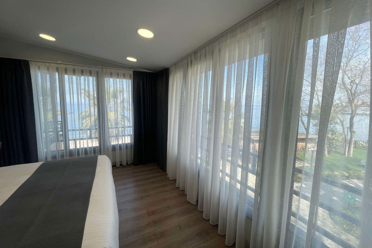 A hotel room with a bed, large windows with sheer curtains, a view of the outdoors, and recessed ceiling lights.