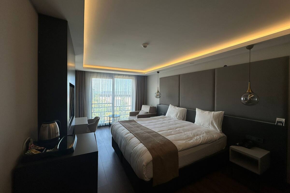 A modern hotel room with a large bed, bedside tables, a desk with a chair, and a view through the window.