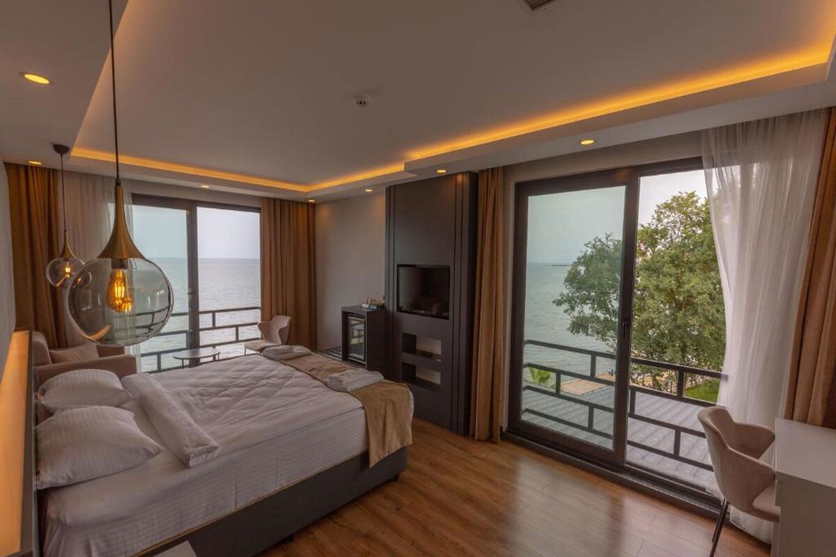 A modern bedroom with a large bed, a TV, and a balcony with a view of the sea.