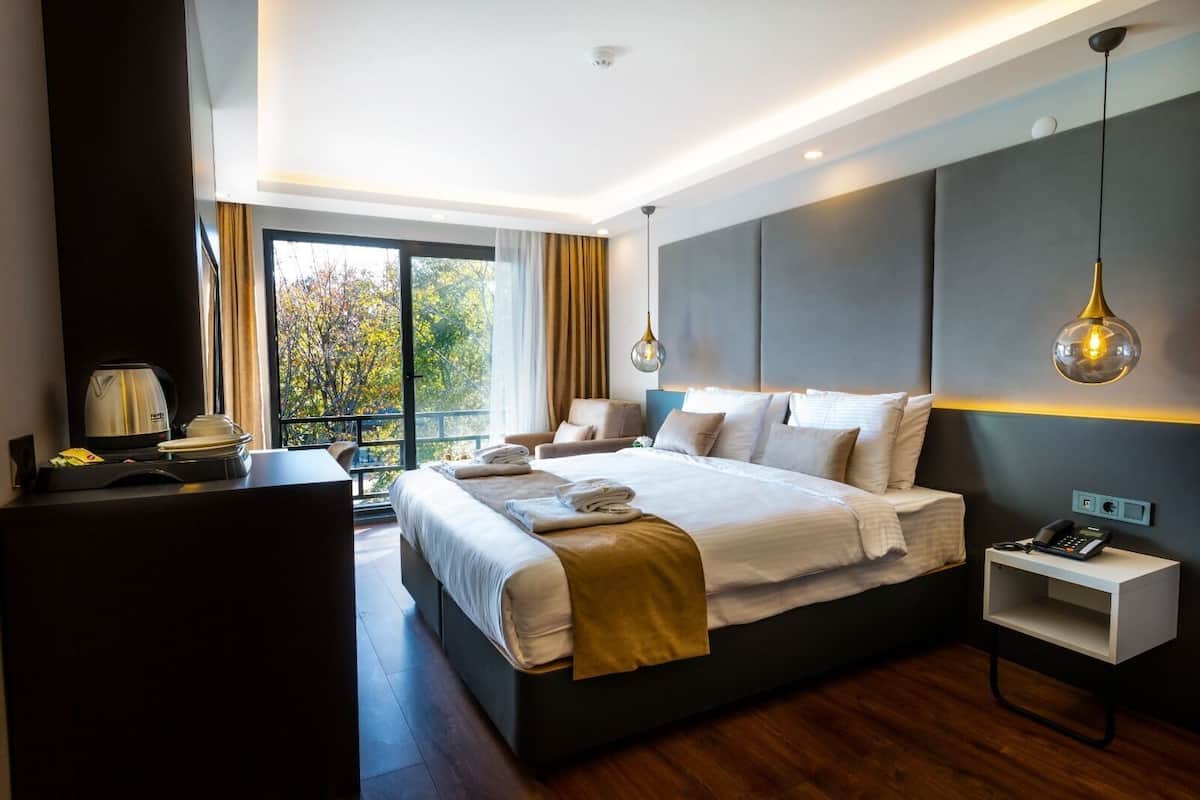 A modern hotel room with a large bed, bedside tables, a desk, and a view of trees through the window.