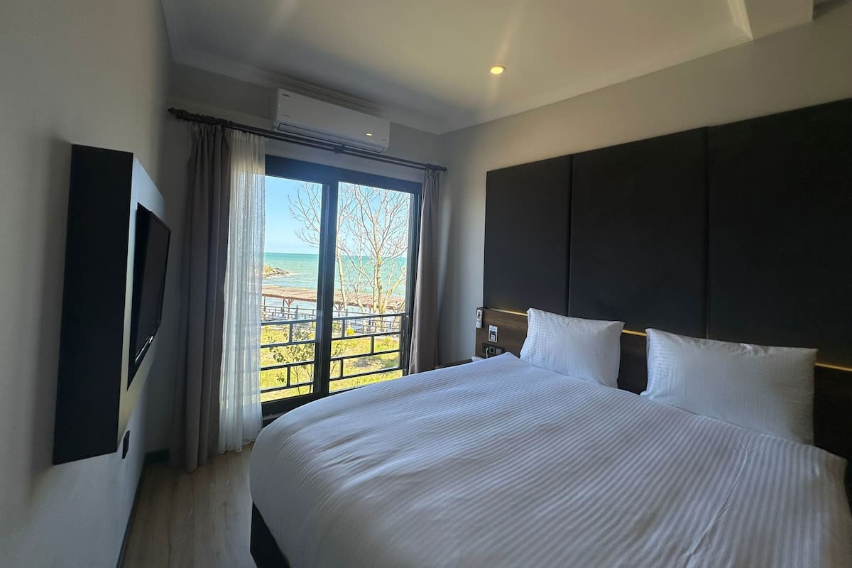 A hotel room with a large bed, a flat-screen TV, an air conditioning unit, and a balcony with a view of the sea.