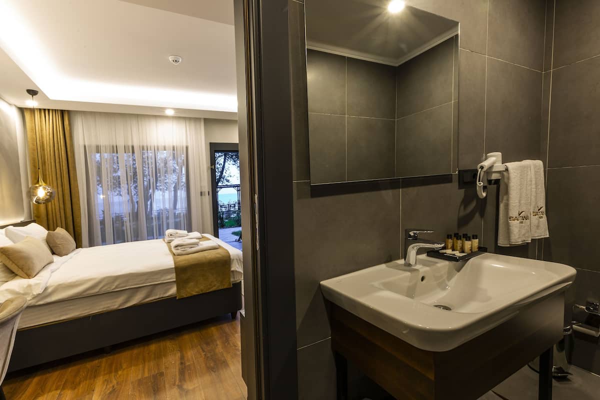 A modern hotel room with a bed, a bathroom with a sink and mirror, and a view of the outdoors through the window.