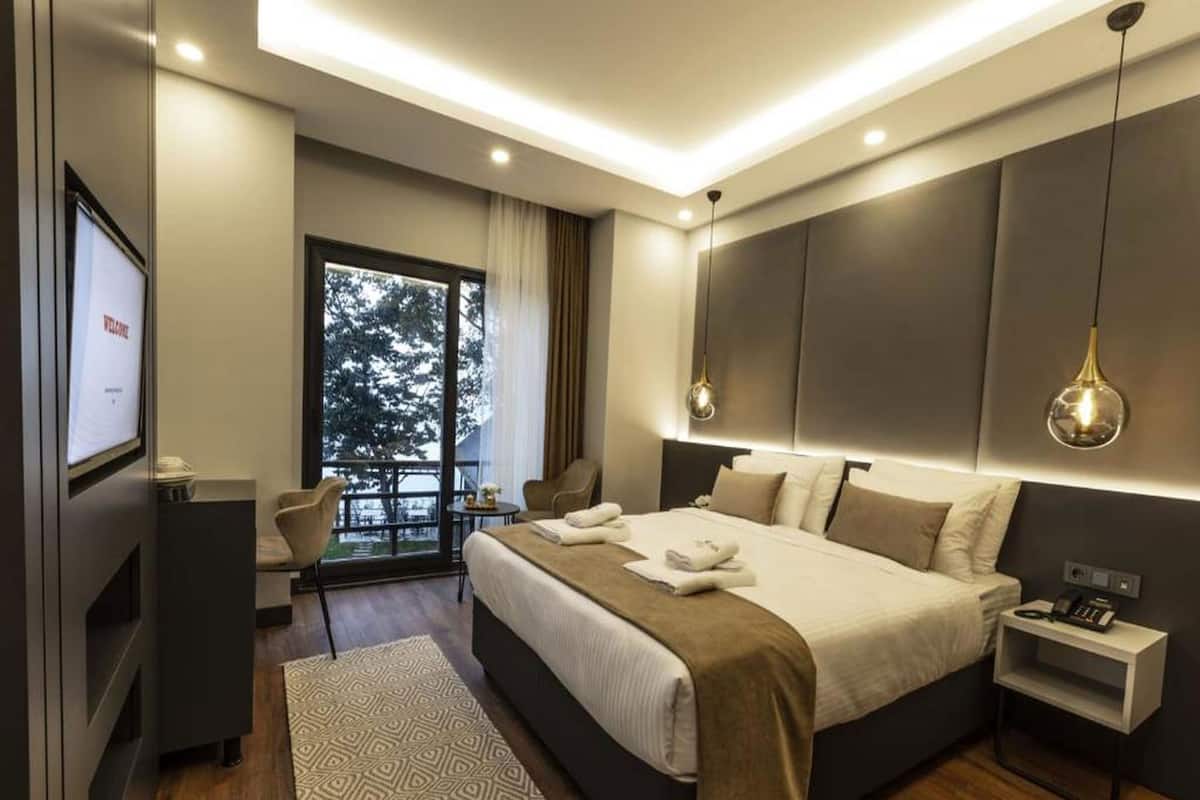 A modern hotel room with a large bed, bedside tables, a TV, and a balcony with a view.