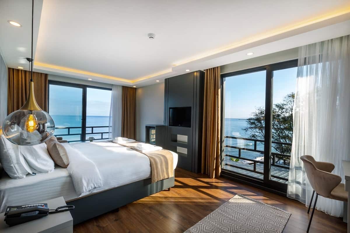 A modern bedroom with a large bed, a TV, and a balcony with a view of the sea.
