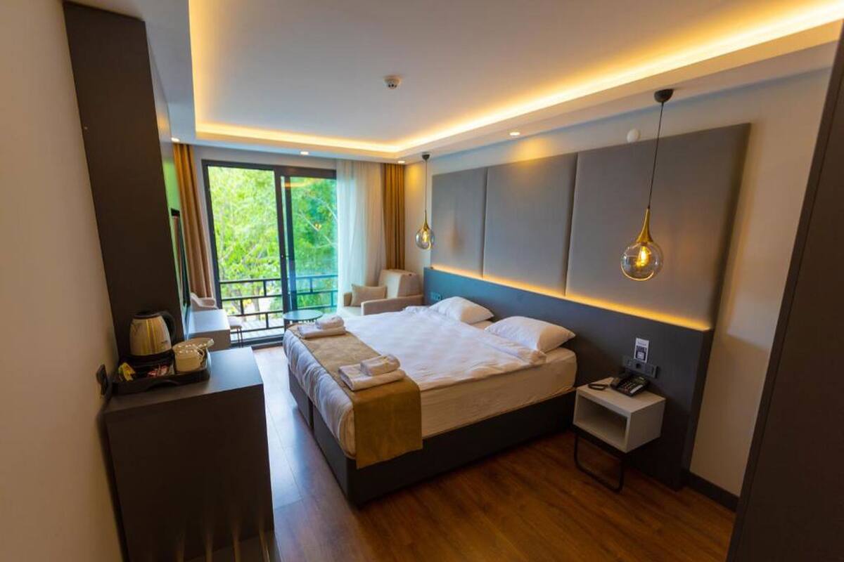 A modern hotel room with a large bed, a small desk, a nightstand, and a view of greenery outside.