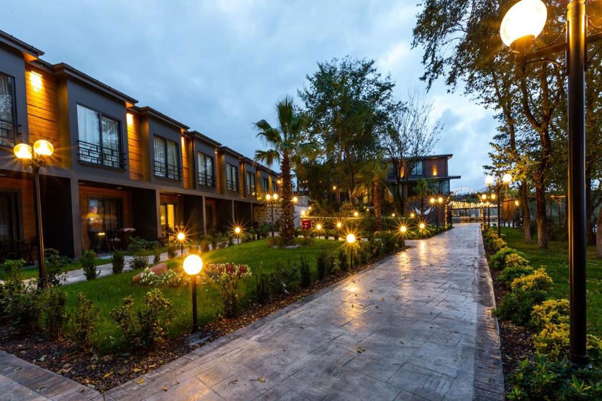 A pathway lined with evenly spaced street lamps, leading to a row of modern buildings with large windows and a well-maintained garden area.