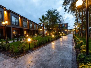 Un chemin bordé de lampadaires régulièrement espacés, qui mène à une rangée de bâtiments modernes dotés de grandes fenêtres et agrémenté d’un jardin bien entretenu.