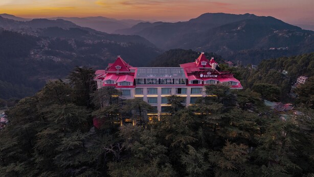 Un edificio grande de varios pisos con techo rojo, rodeado de un bosque denso y montañas al fondo.