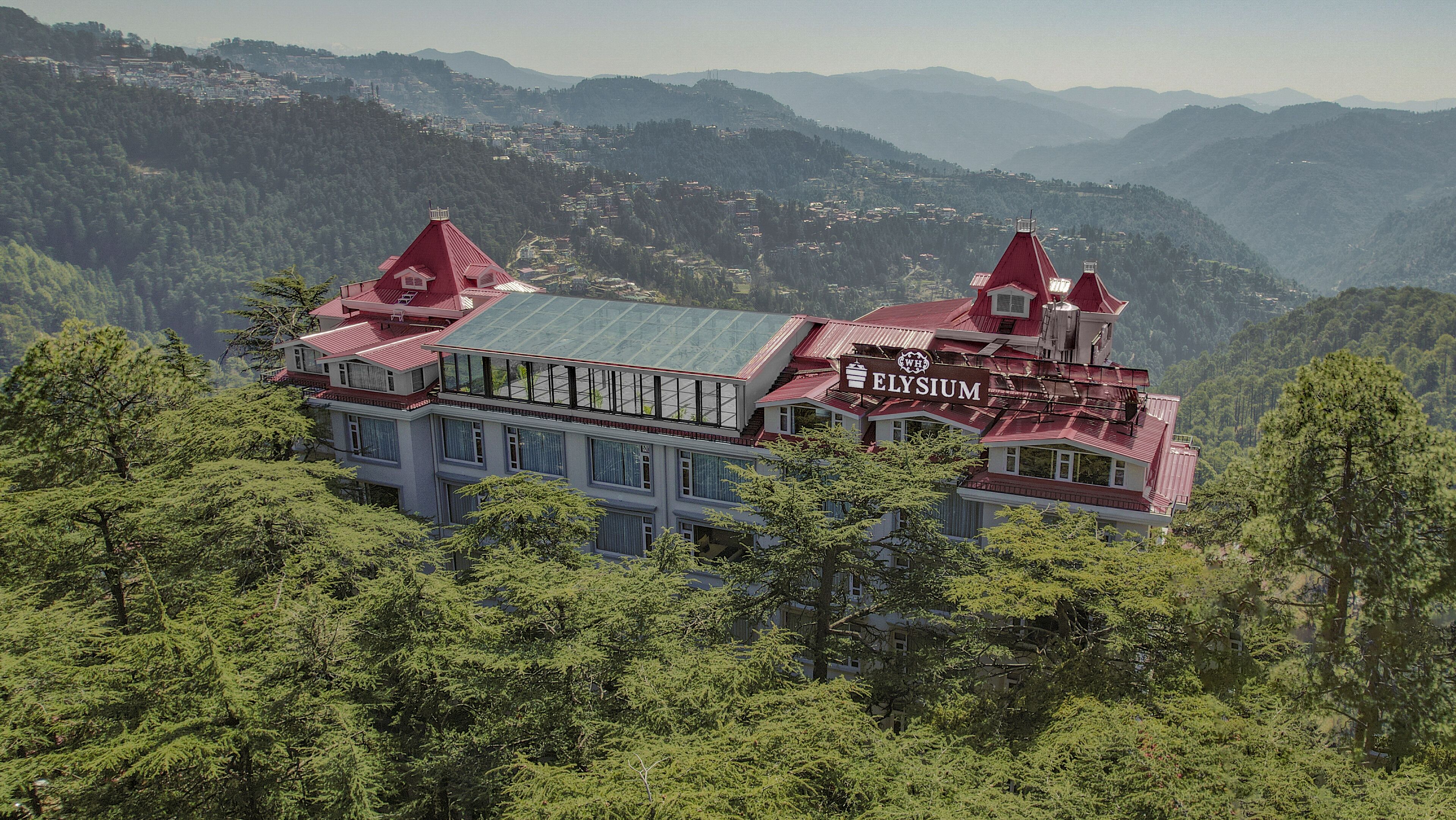 A large, multi-story building with a red roof and a sign that reads "ELYSIUM." It is surrounded by dense green forest and mountains in the background.