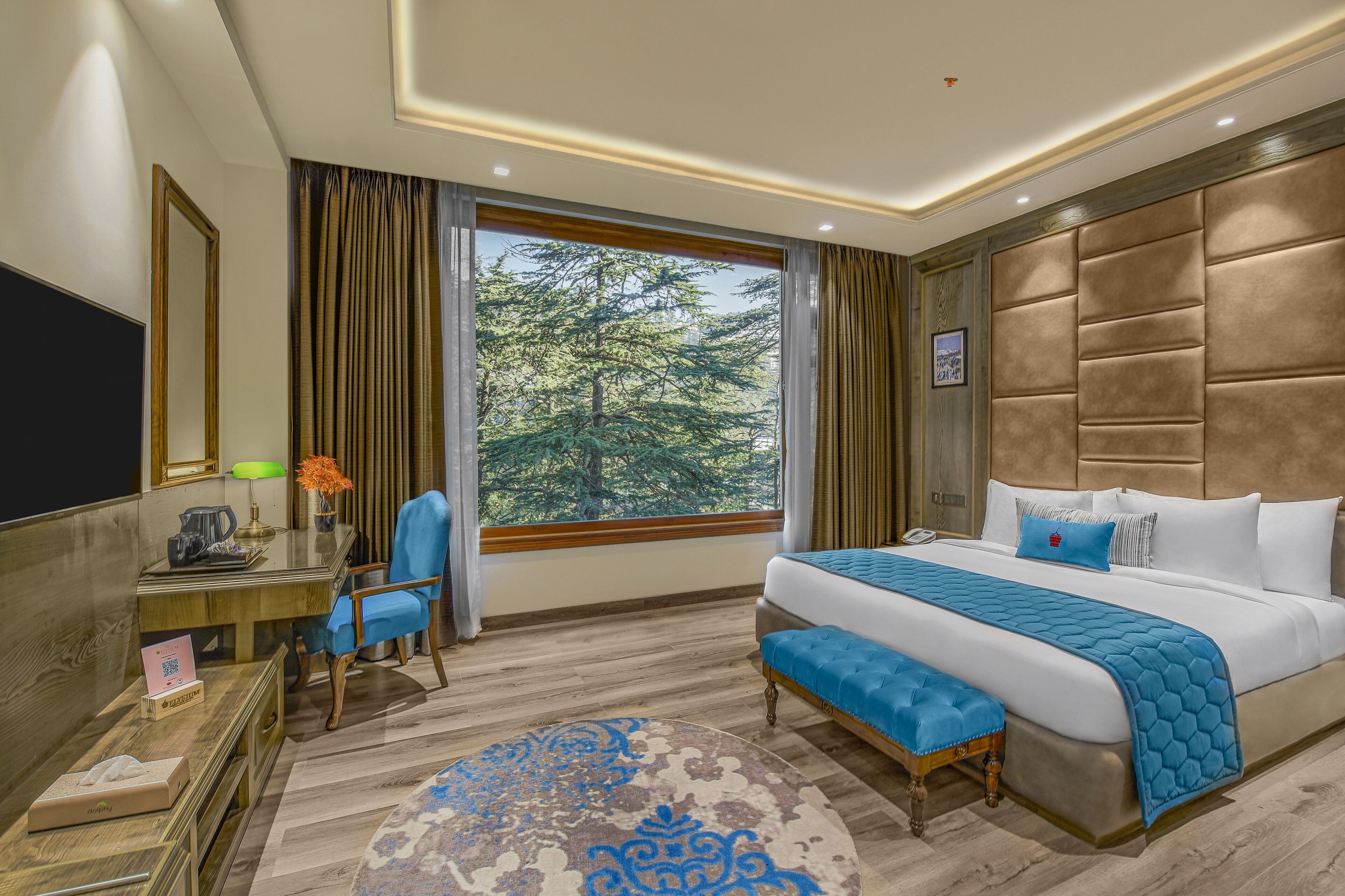 A hotel room with a large bed, a desk with a chair, a TV, and a window with a view of trees.